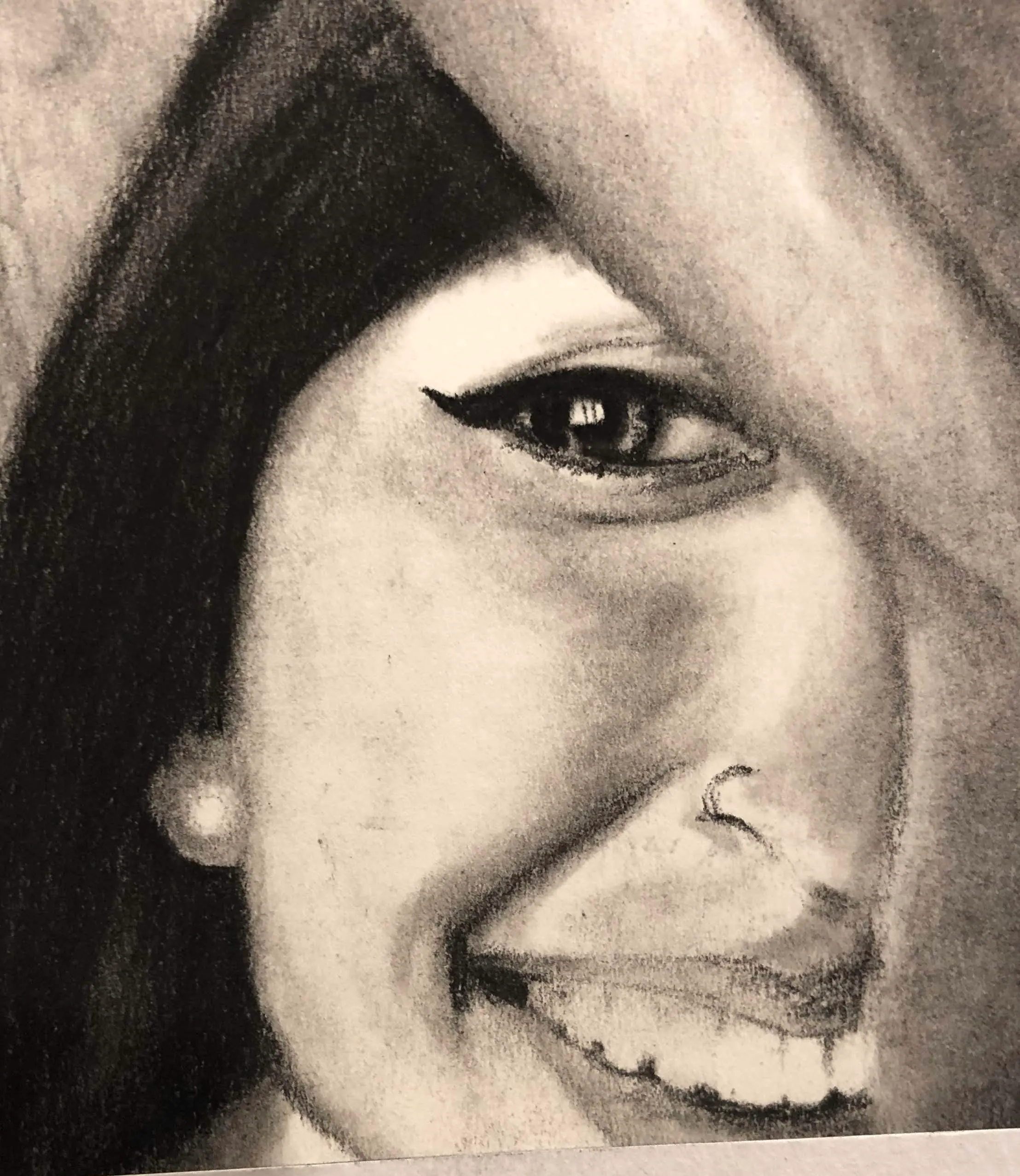 Charcoal pencil close-up of half a woman’s smiling face: one eye with winged liner beneath a fall of dark hair, teeth showing.