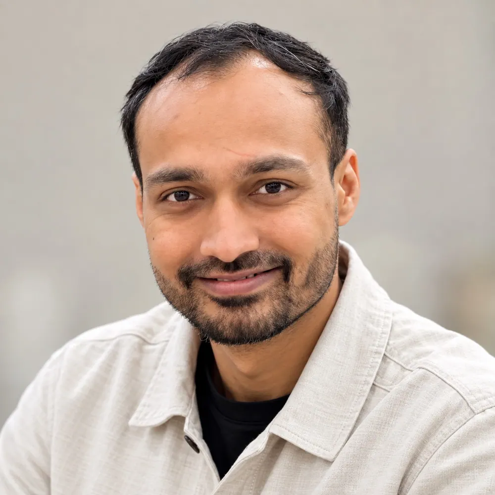 Photograph of the artist: a smiling man with short dark hair and a short beard, in a light linen-textured shirt over a black t-shirt, against a soft grey background.